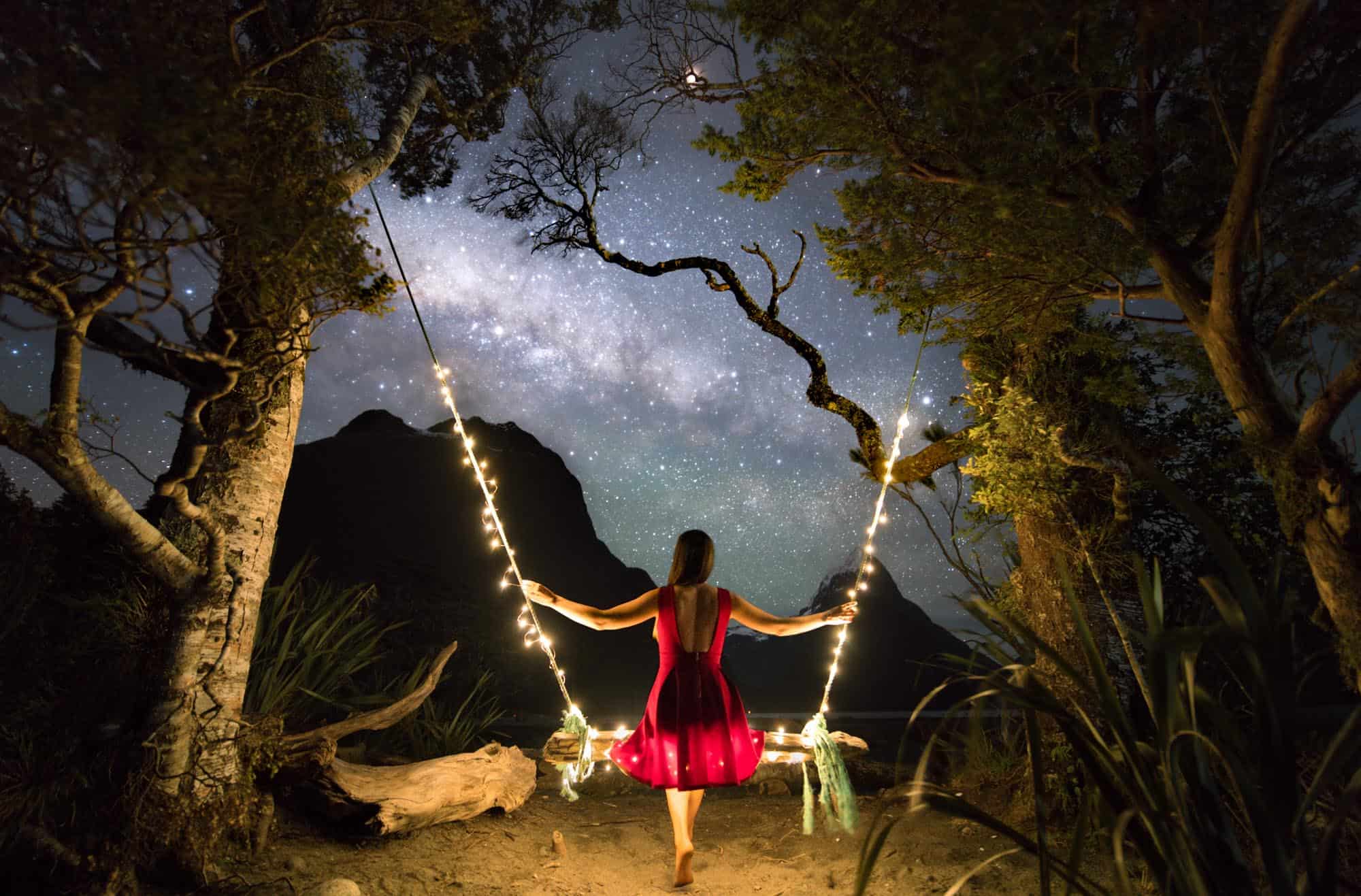A girl in a red dress sat on a swing surrounded by fairy lights looking out towards the milky way over Milford Sound in New Zealand