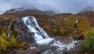 The Meeting of the Three Waters viewpoint near Glencoe shows a larger waterfall on the left and smaller waterfall on the right with mountains and low hanging clouds in the background.