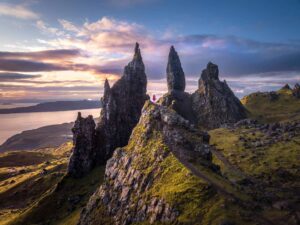 A young woman in a red dress appears as a small silhouette between two large pillars of rock with a purple sunrise sky at Old Man of Storr. With 10 Days in Scotland, sunrise at the Old Man of Storr is a must.