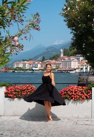 A girl twirls in front of pretty red flowers that decorate the lakeside promenade with stunning views back towards Bellagio town centre.