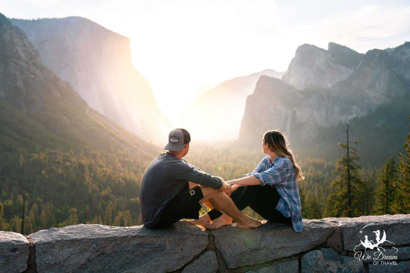 A couple sat on a wall holding hands while looking out towards Tunnel View at sunrise in Yosemite National Park on an adventurous romantic getaway.