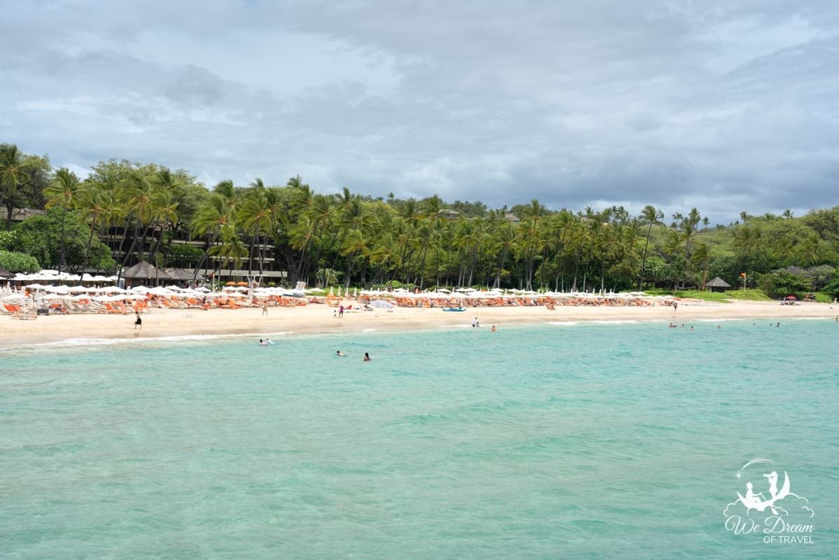 A view of Mauna Kea beach showing the clear water and white sand beach lined with beach chairs and palm trees on the Big Island of Hawaii.