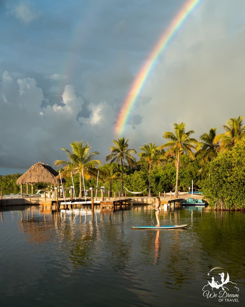 A woman paddleboarding on Key Largo beneath a bright double rainbow with palm trees in the background