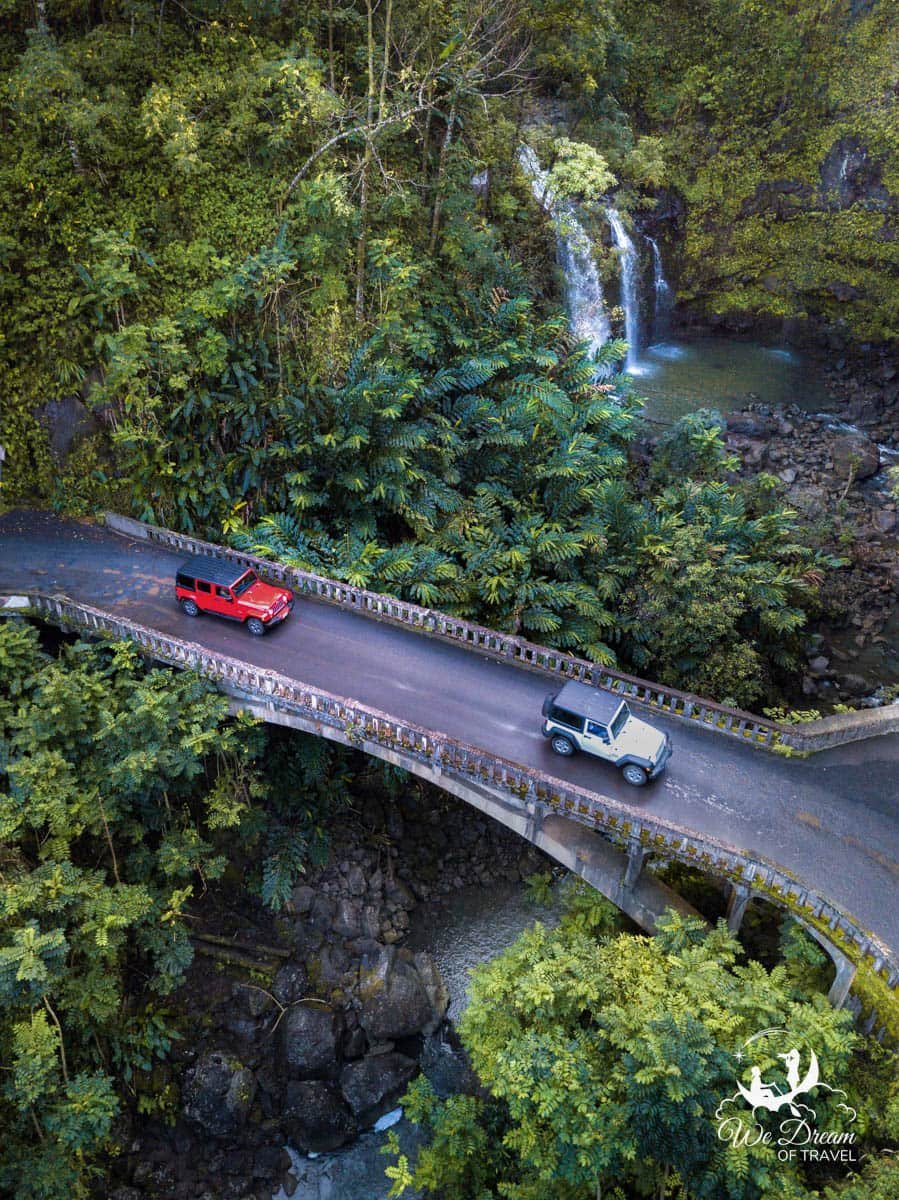 A red and white jeep driving the Road to Hana cross over a bridge with a waterfall in the background.